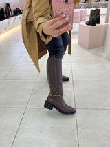 Bota cano longo Beyou Shoes marrom com detalhe de fivela dourada no tornozelo em ambiente de loja