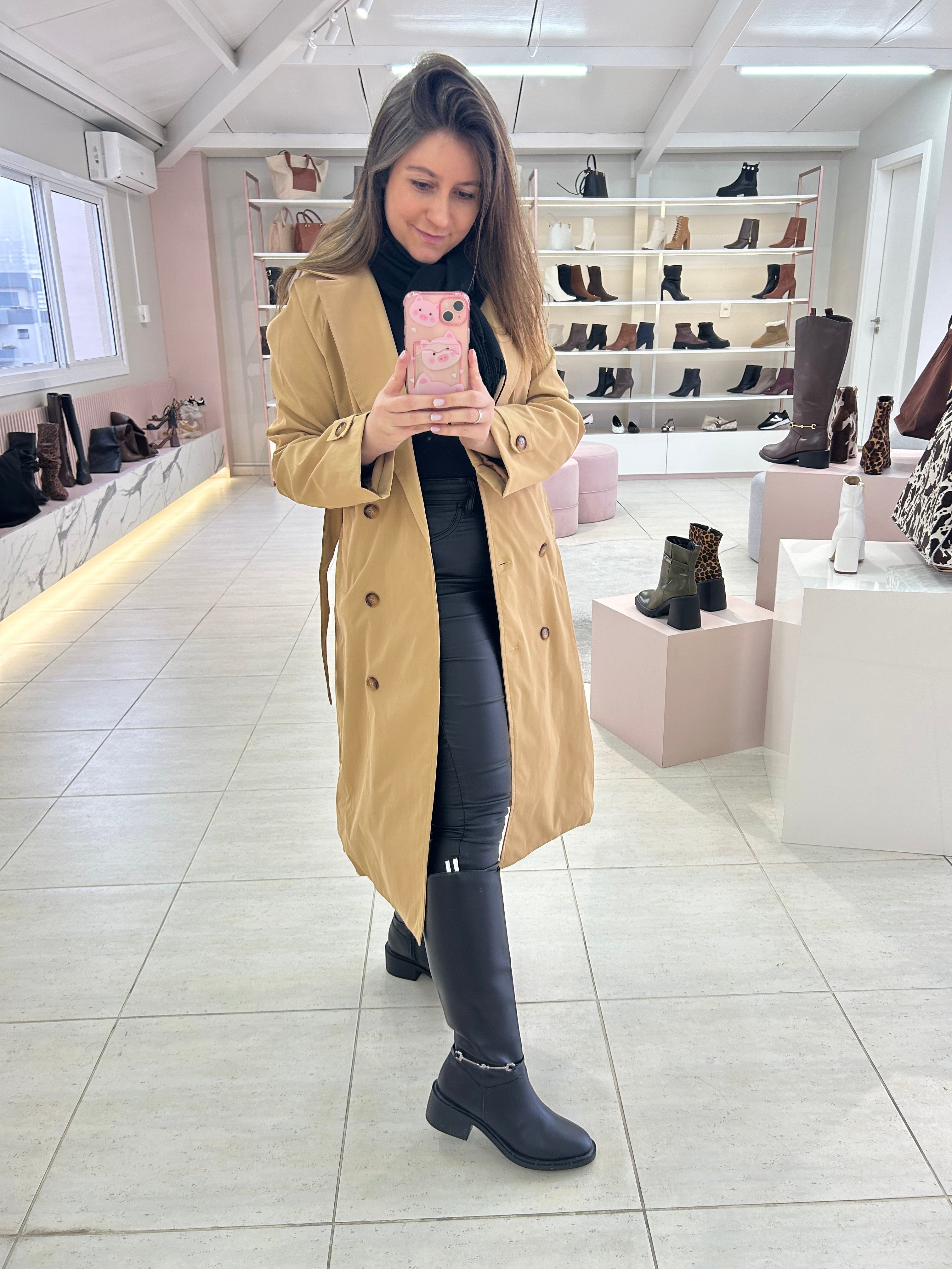 Mulher usando bota cano longo preta Beyou Shoes com trench coat bege em loja de calçados