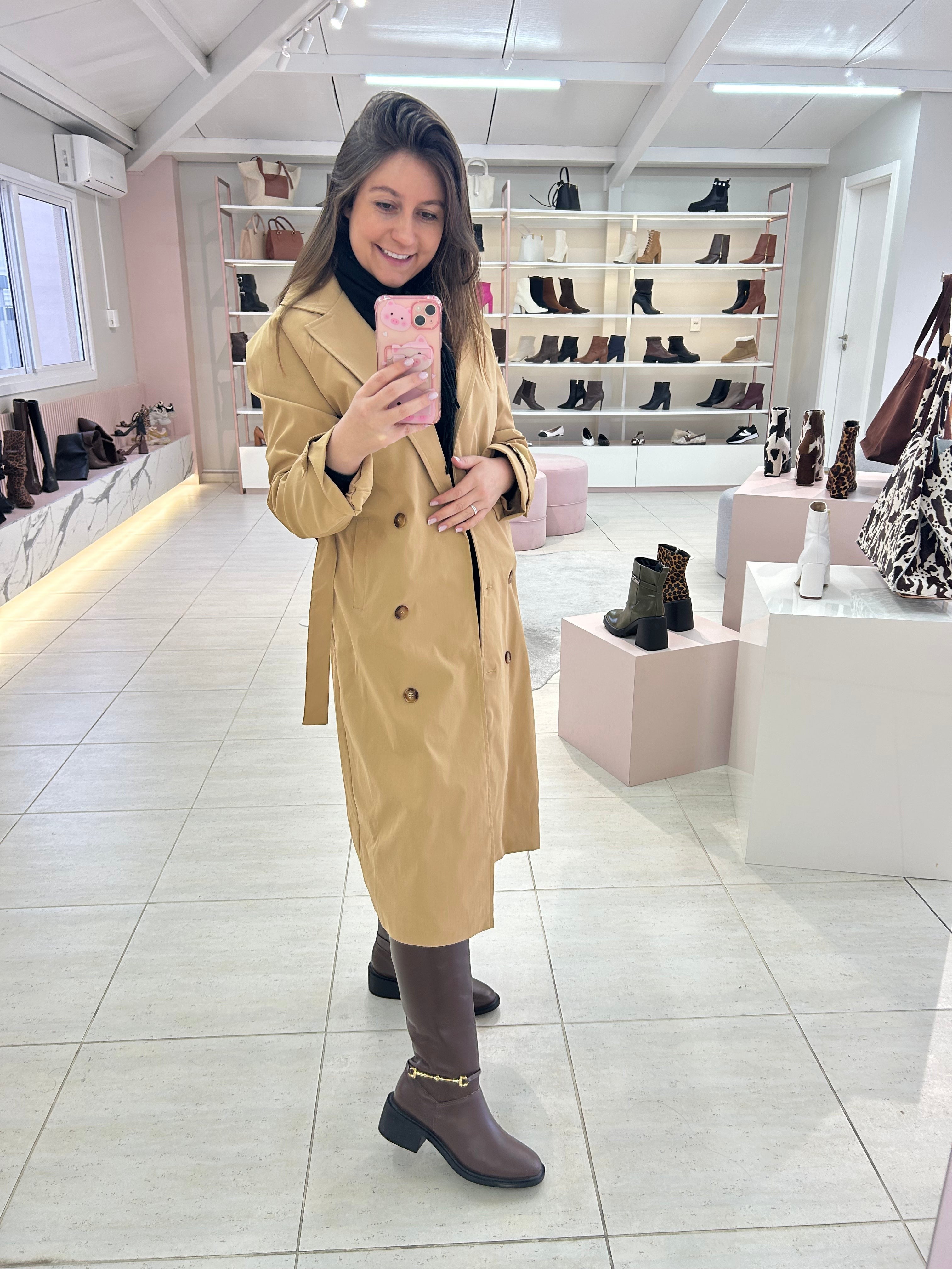 Mulher usando Bota Durazno marrom da Beyou Shoes com trench coat bege em loja de calçados.
