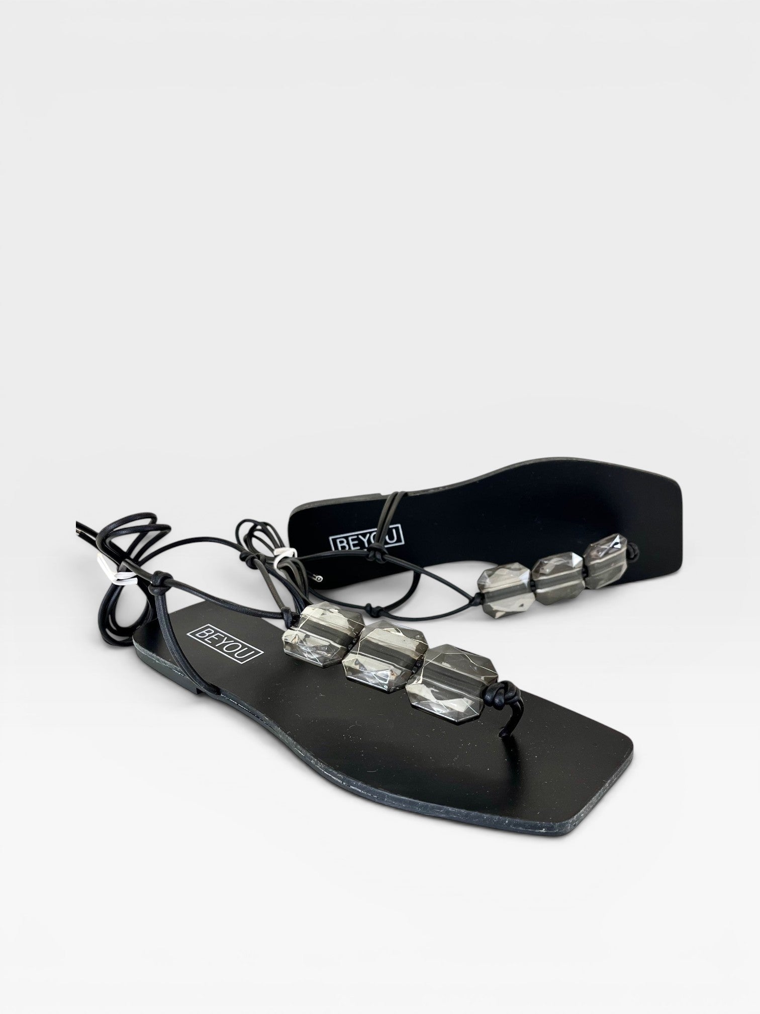 Rasteira preta Beyou Shoes com tiras de amarração e detalhes em pedrarias transparentes sobre fundo branco.