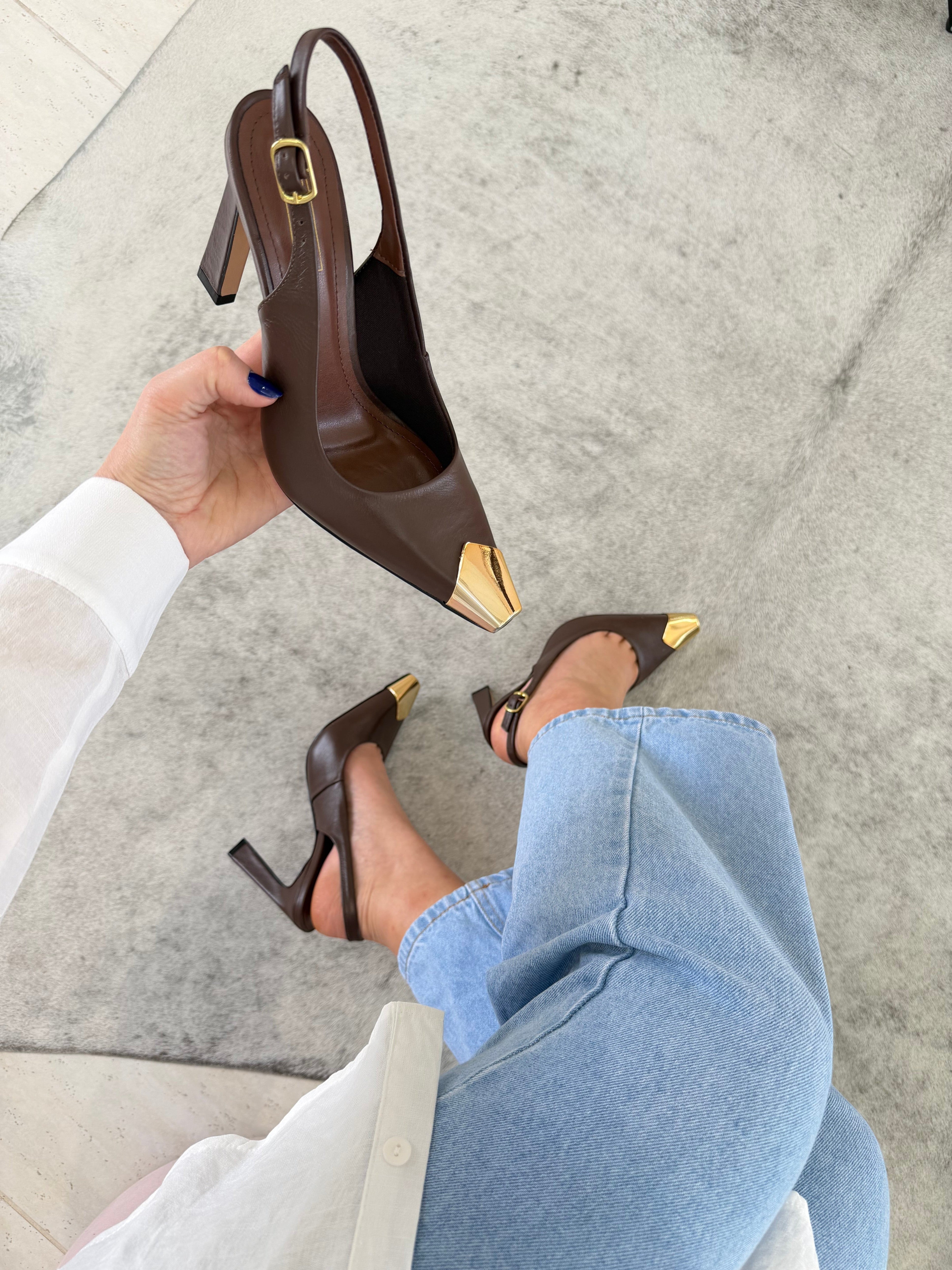 Slingback Beyou Shoes Bristol em couro marrom com ponteira metálica dourada e salto alto, calçado por uma mulher.