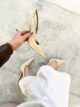 Slingback Beyou Shoes off white em couro com bico fino e salto taça, sendo segurado por uma mão e calçado no pé.